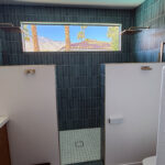Walk-in shower with vertical dark teal midcentury tile, dual rainfall showerheads, privacy divider walls, and a walnut double vanity in a newly added ensuite bathroom.