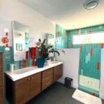 Midcentury-style bathroom with floating walnut double vanity, vertical teal and orange shower tile, dual mirrors, and natural light from clerestory window.