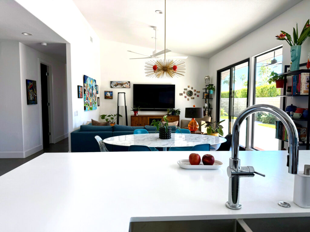 Open-plan den with large sliding glass doors, added windows for mountain views, white island counter in foreground, and midcentury modern furnishings.