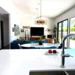 Open-plan den with large sliding glass doors, added windows for mountain views, white island counter in foreground, and midcentury modern furnishings.